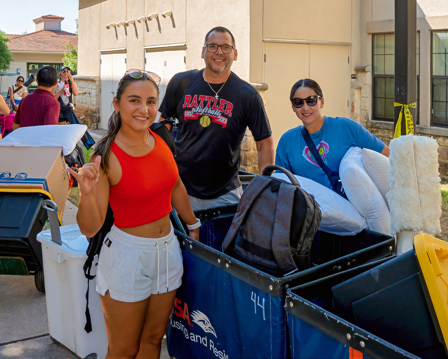 From left to right, Three people with a cart containing bags for move in, A woman smiling holding the schools "birds up" hand gesture, a man smiling, a woman with sunglasses smiling.